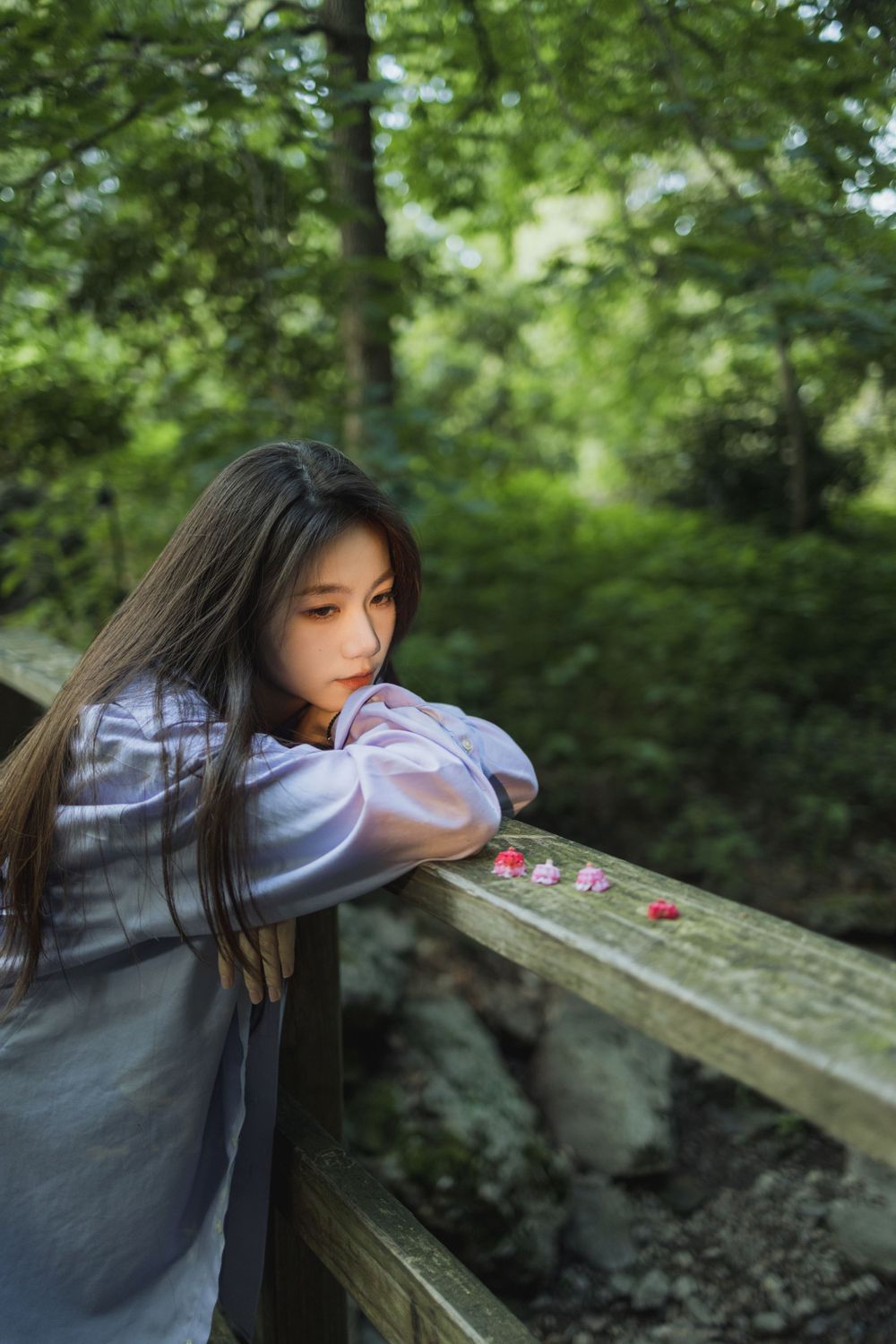 Person in lavender dress leans thoughtfully on wooden railing in lush forest setting.