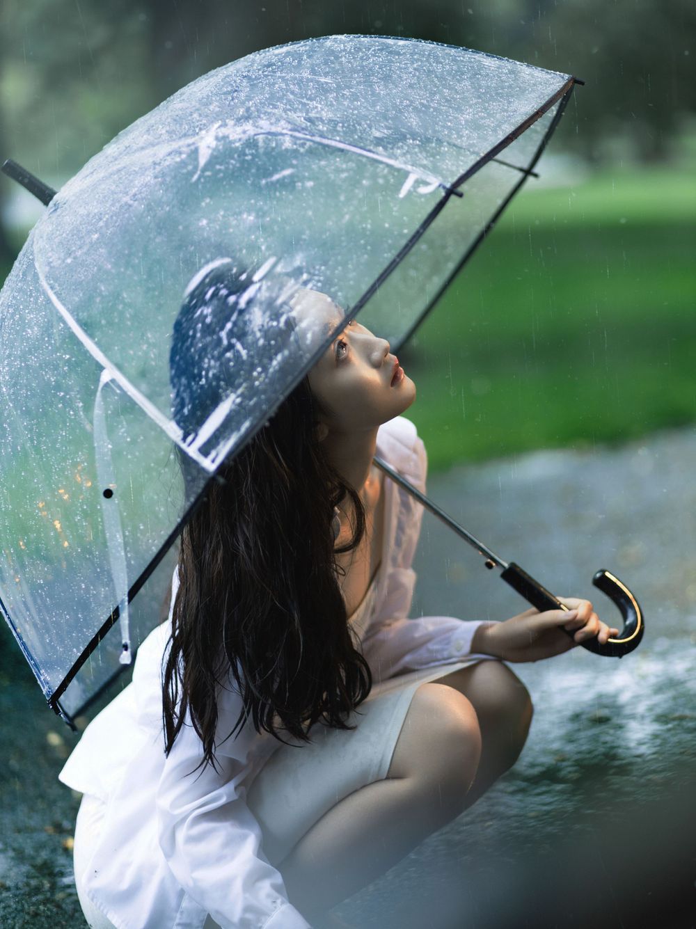 Clear umbrella creates dreamy effect during rainy photoshoot in park setting.