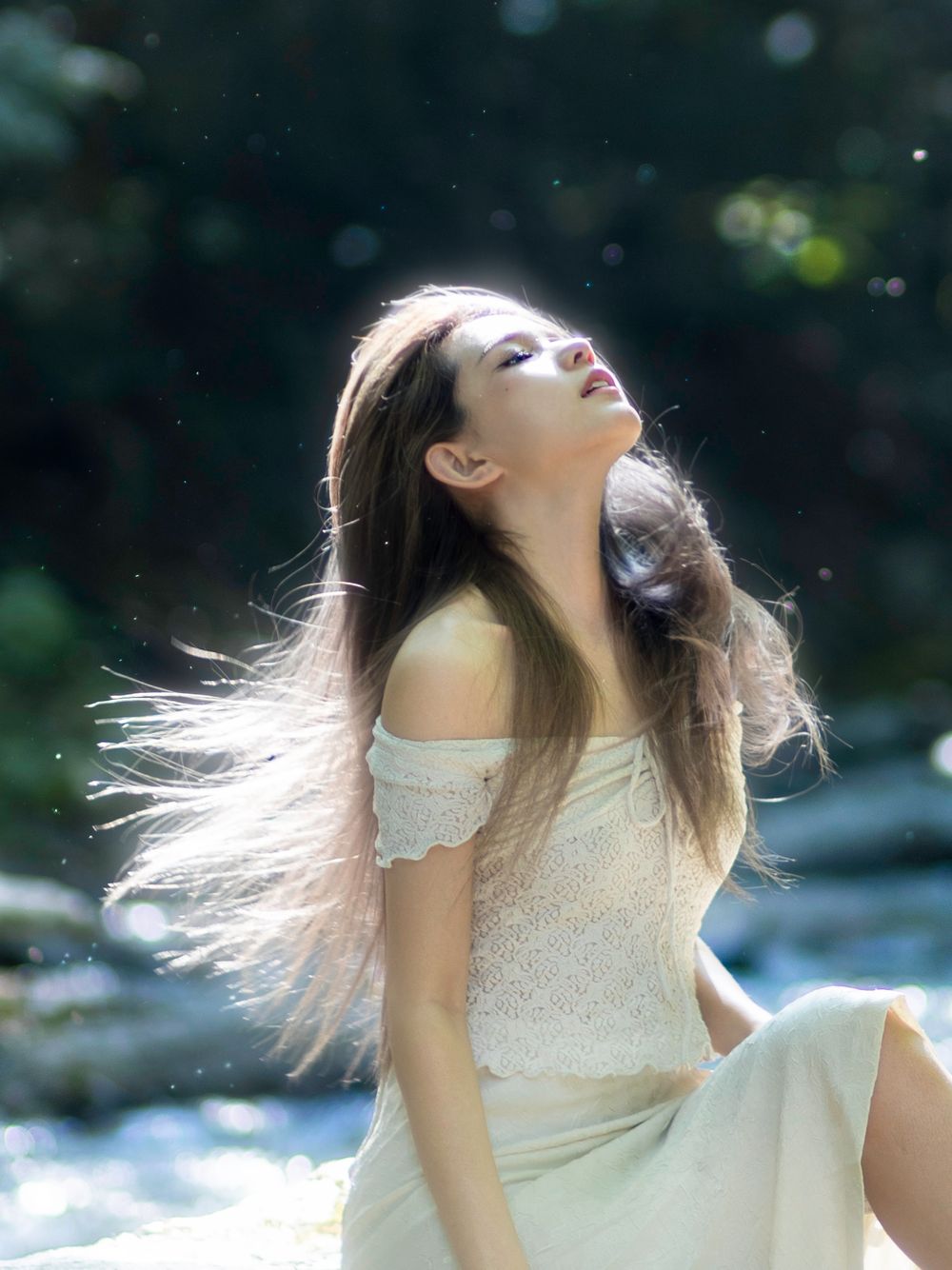 Woman in white dress with flowing hair poses in sunlit nature setting.