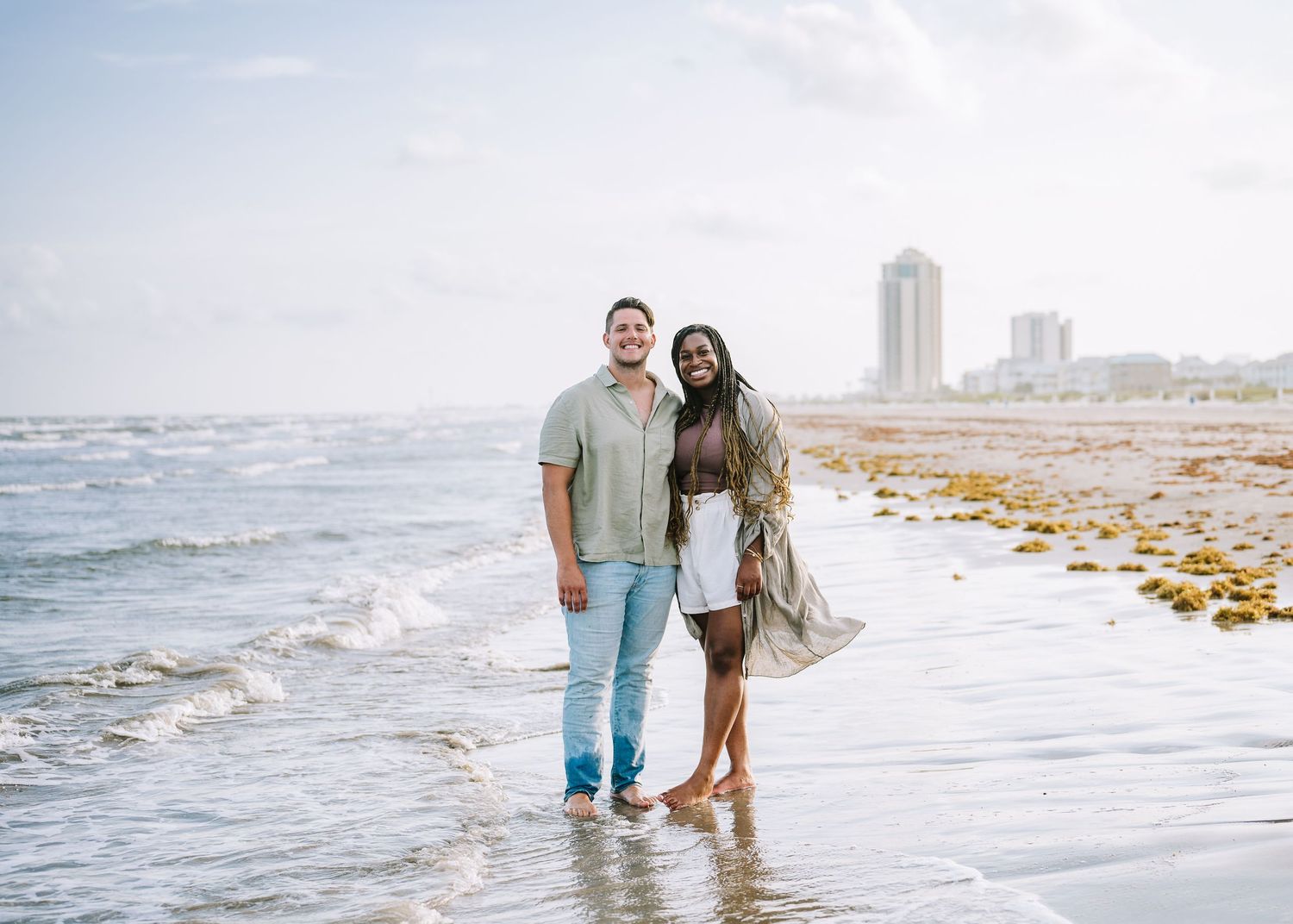 Beach engagement session with couple walking through waves as city skyline appears in background.