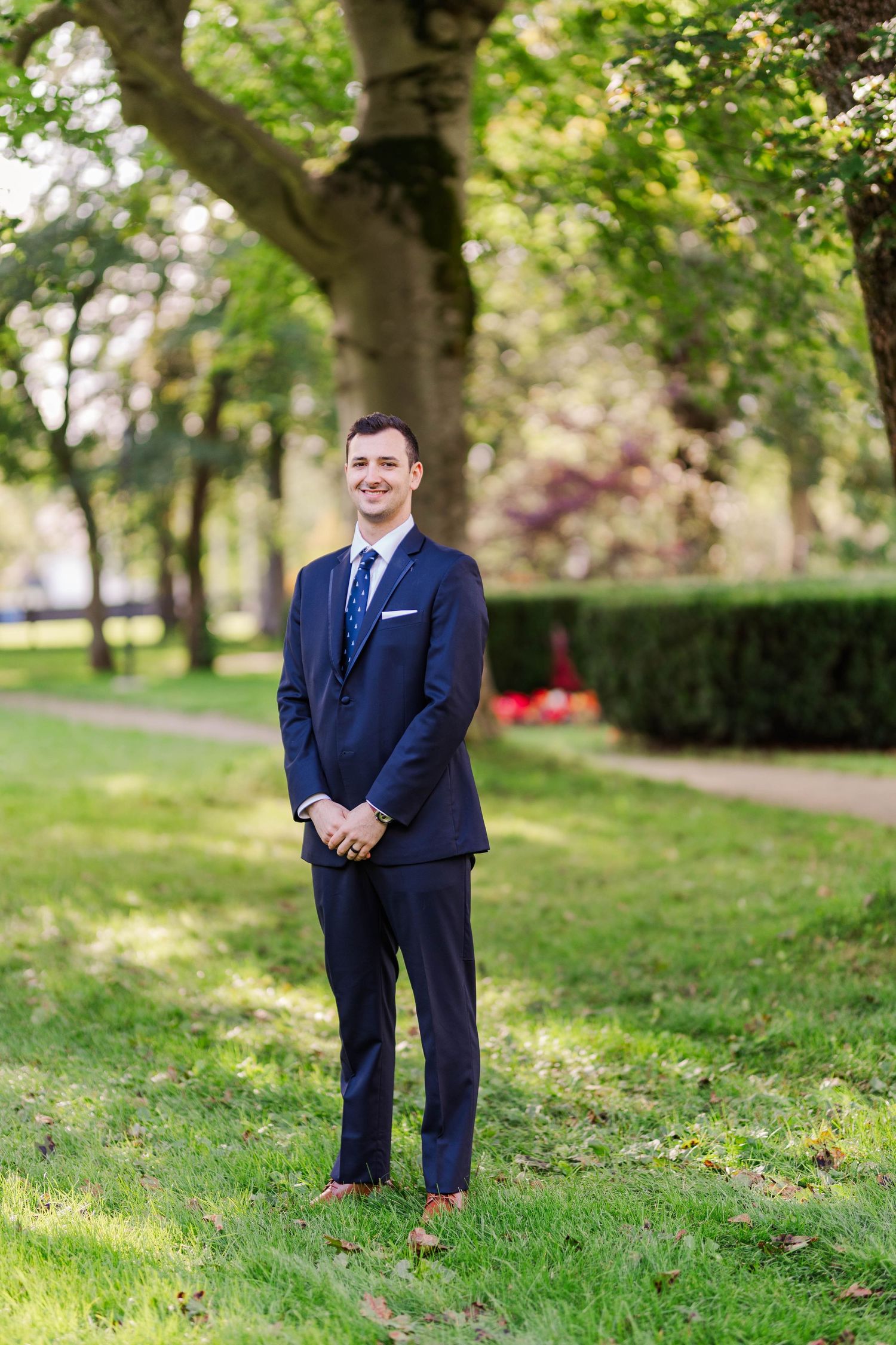 A groom in a navy suit stands on manicured lawn with trees in background.