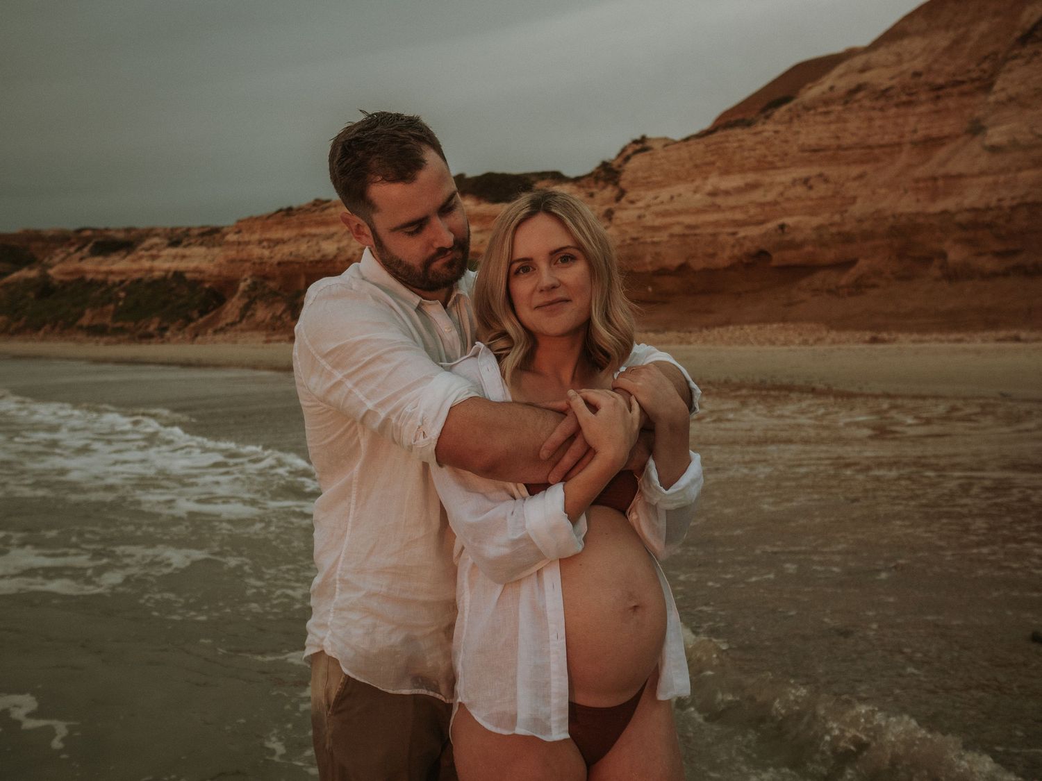 Maternity photoshoot of couple embracing on beach with red cliffs at sunset.
