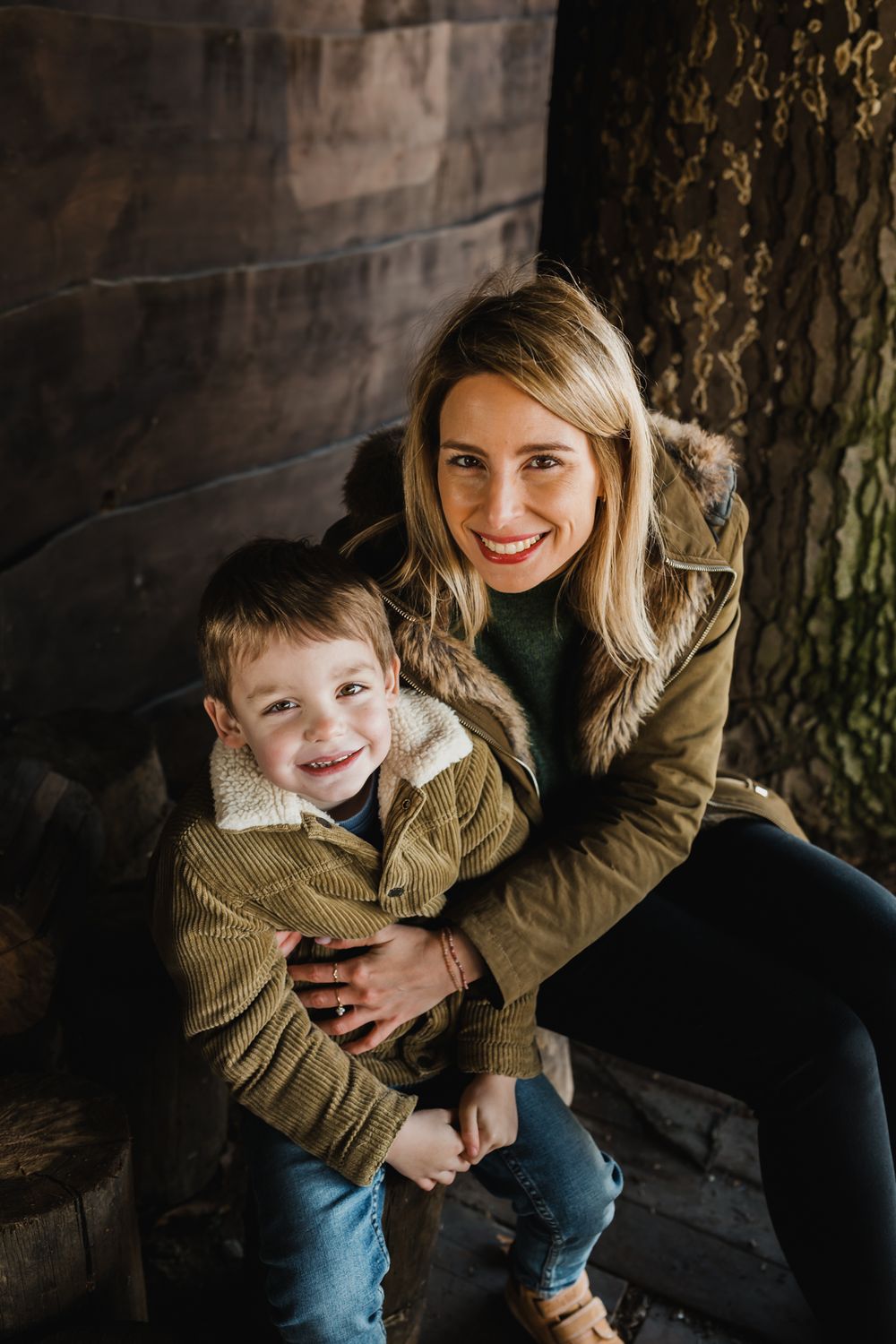 A warm candid portrait of a parent and child sitting together in matching brown jackets against a rustic wooden wall.