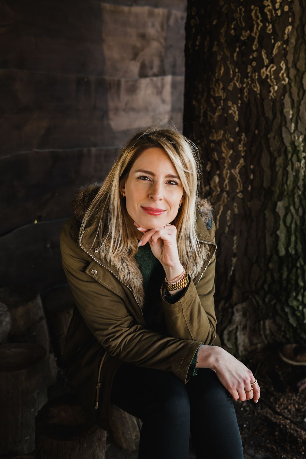 A casual portrait of someone in an olive jacket sitting against a textured wooden wall.