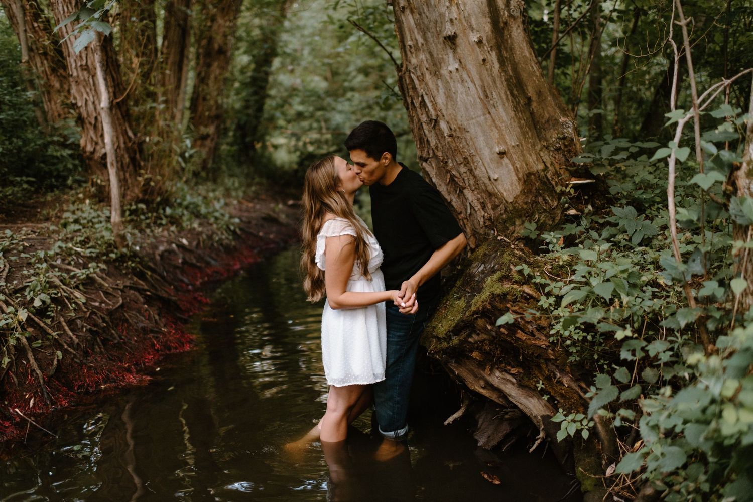 Couple shares intimate moment standing in creek water surrounded by lush forest scenery.