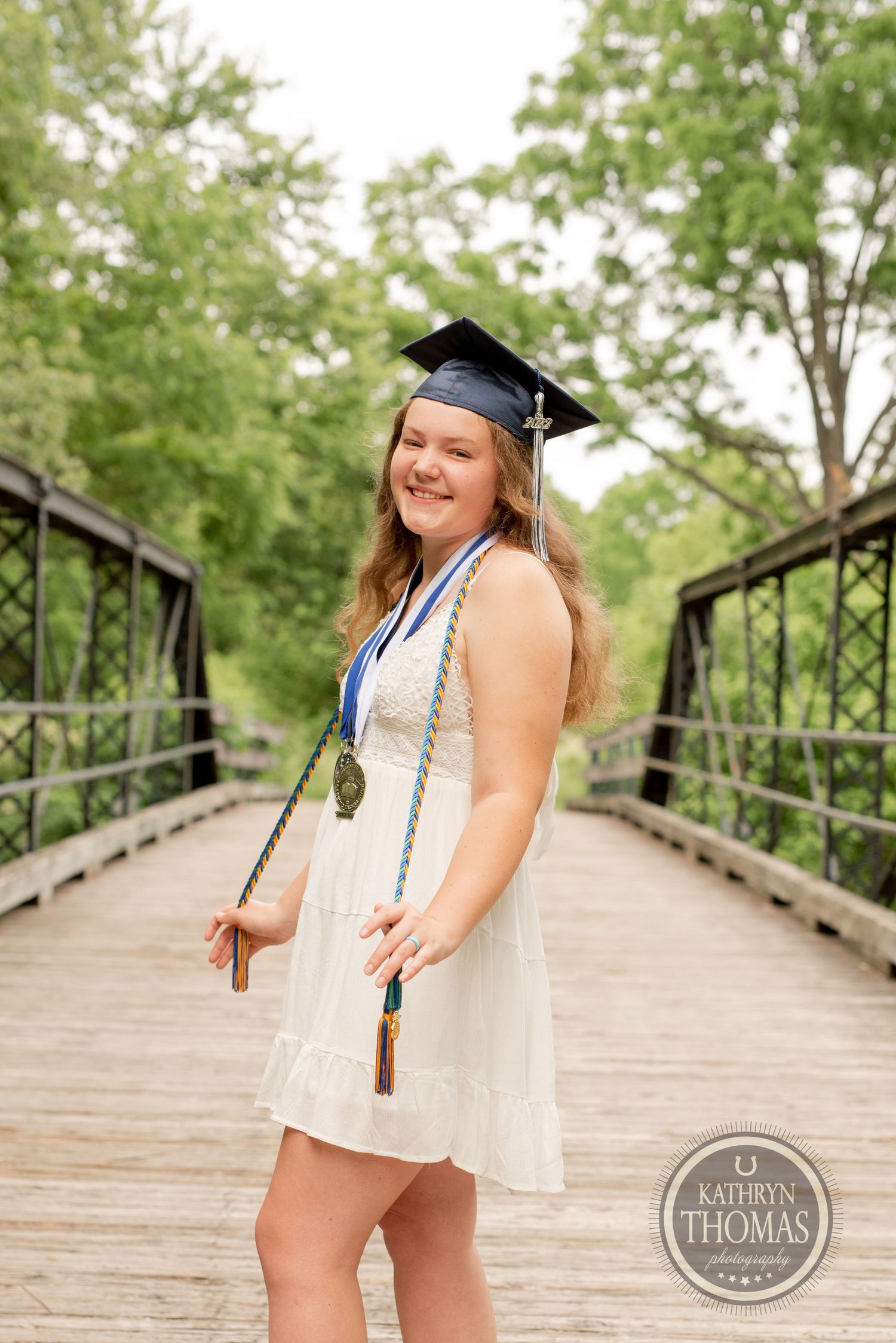 A graduate in white dress and cap poses with honors cords on a rustic bridge.
