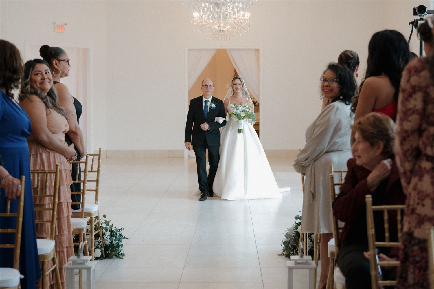 A wedding ceremony in progress inside a bright white venue with guests seated in rows and floral aisle decorations.