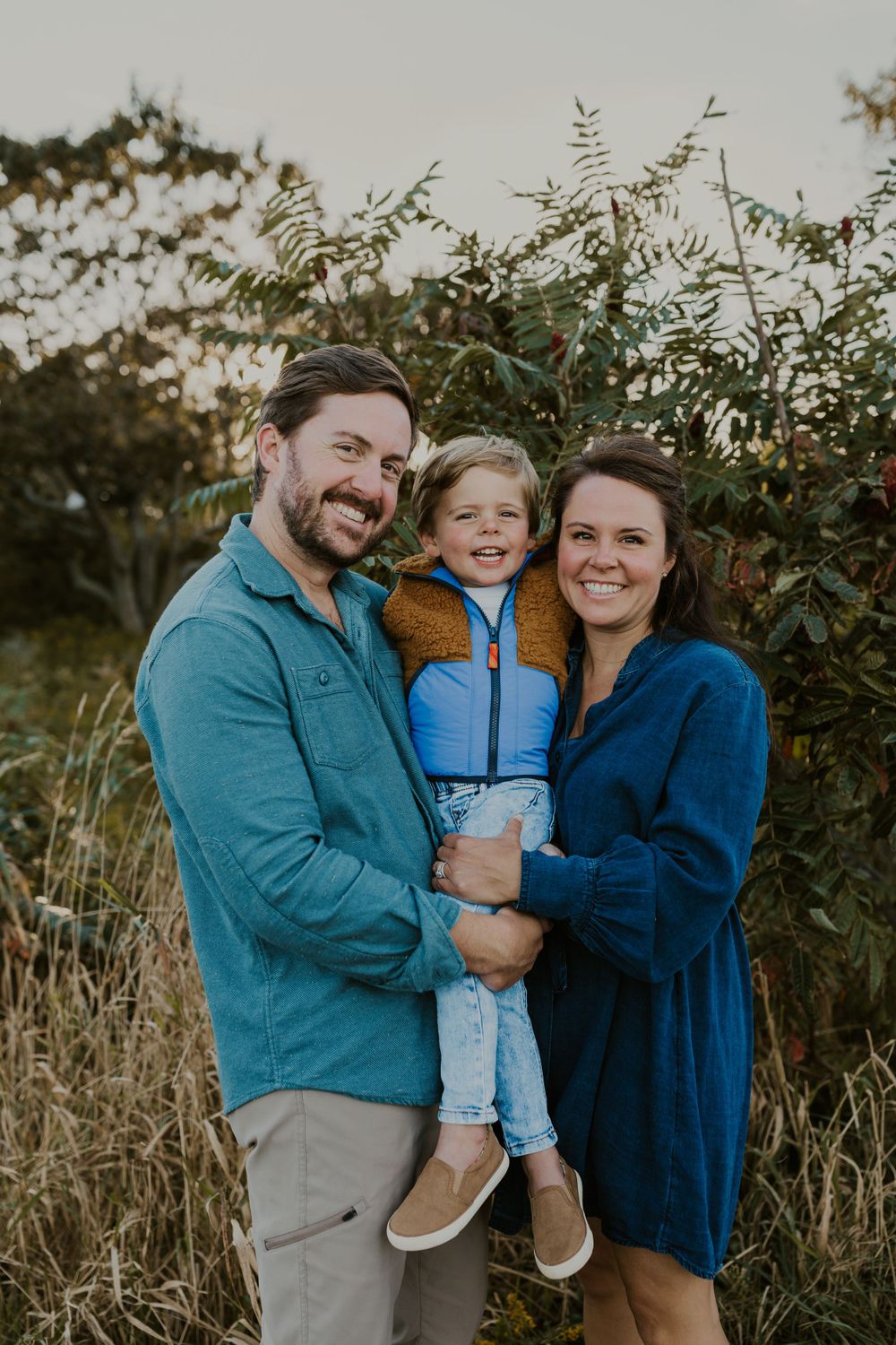 Young family of three smiling together against a natural autumn backdrop.