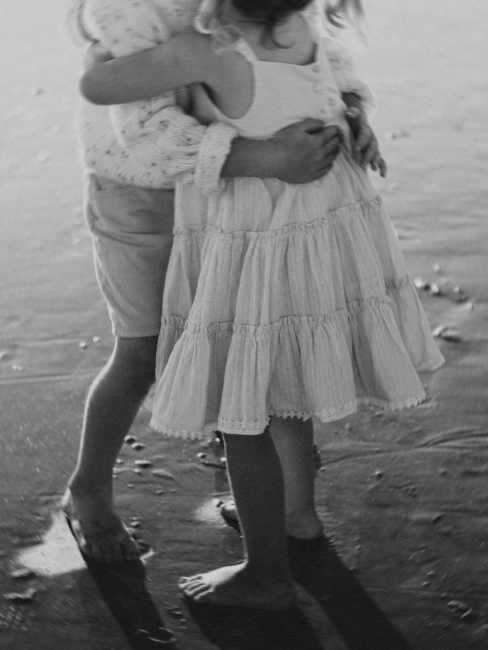 Two young children in summer clothes standing barefoot on a beach in a black and white photograph.