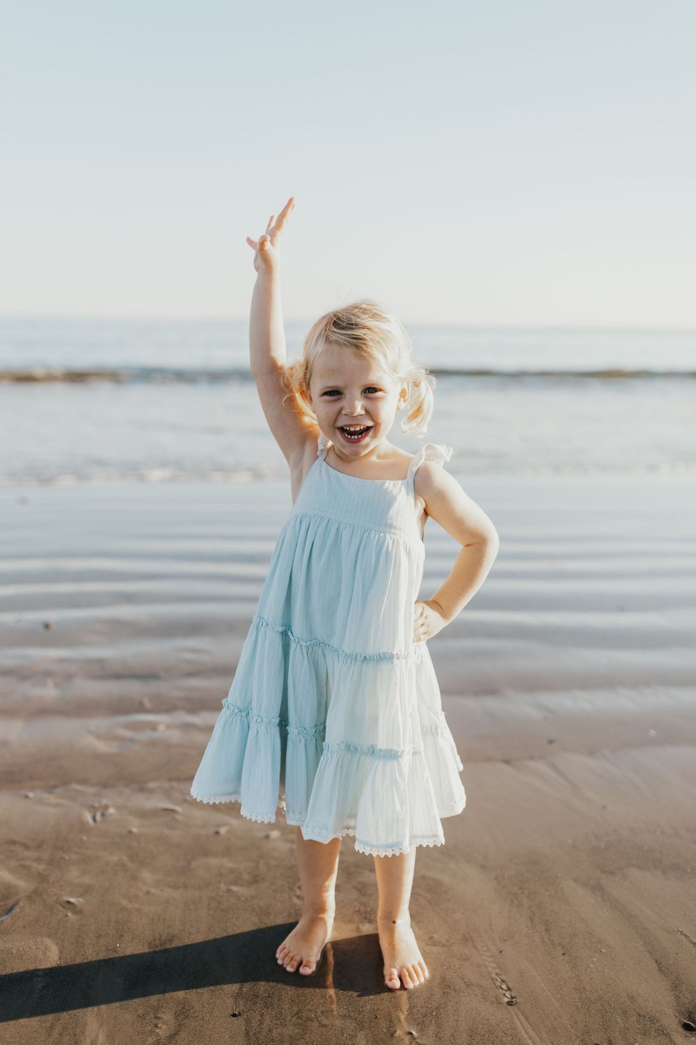 Young child in a light blue tiered dress striking a playful pose on the beach.