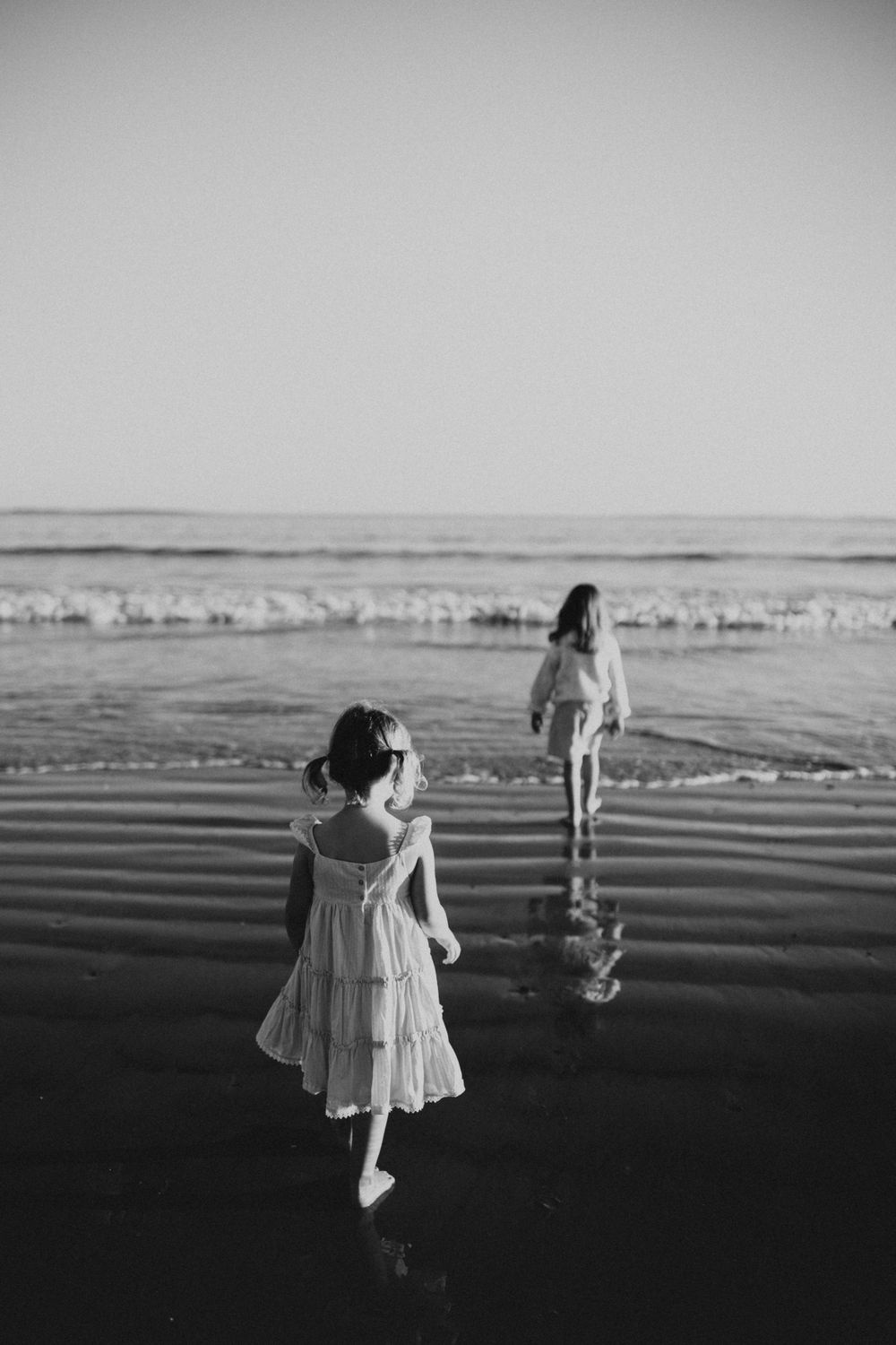 Black and white photo of two children in dresses walking through shallow ocean water.
