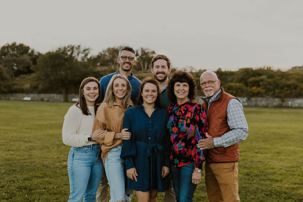 Multi-generational group poses outdoors on grassy field at sunset.