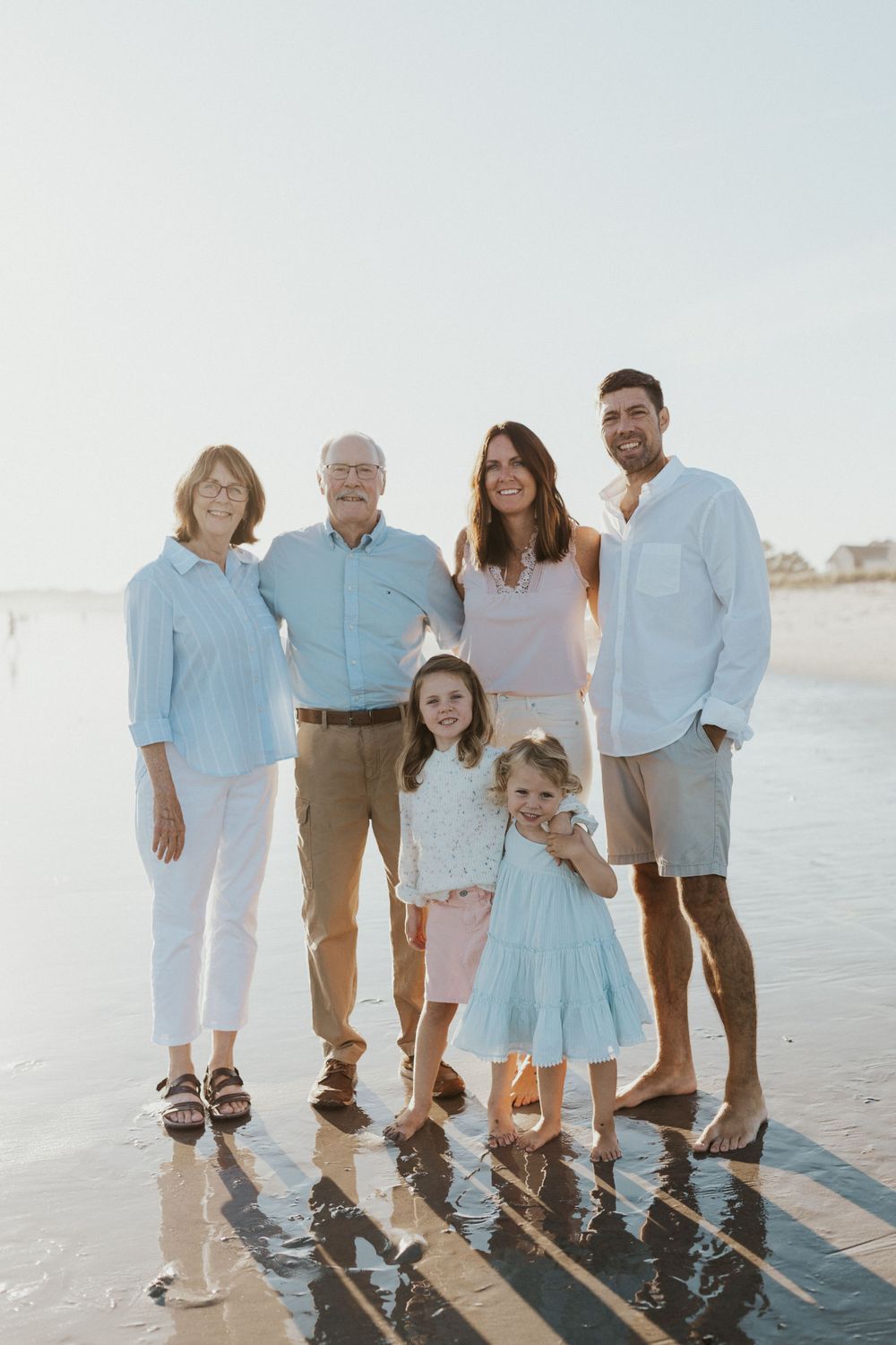 Multi-generational family portrait on beach with long shadows cast on wet sand at sunset.