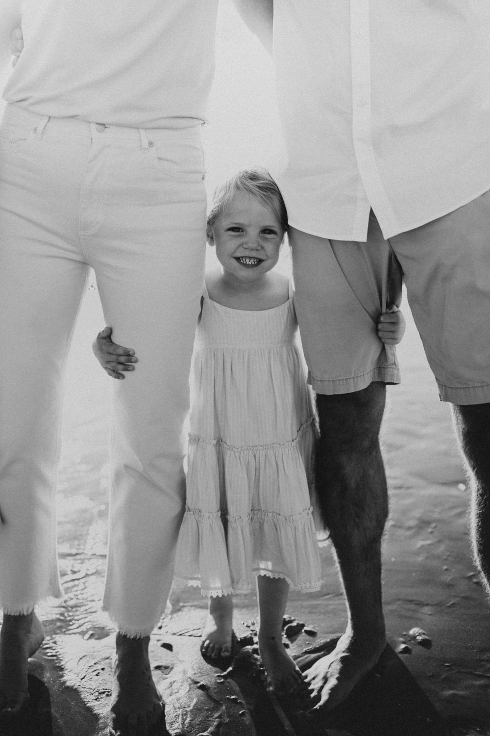 Small child in a white tiered dress standing between two adults on a beach.