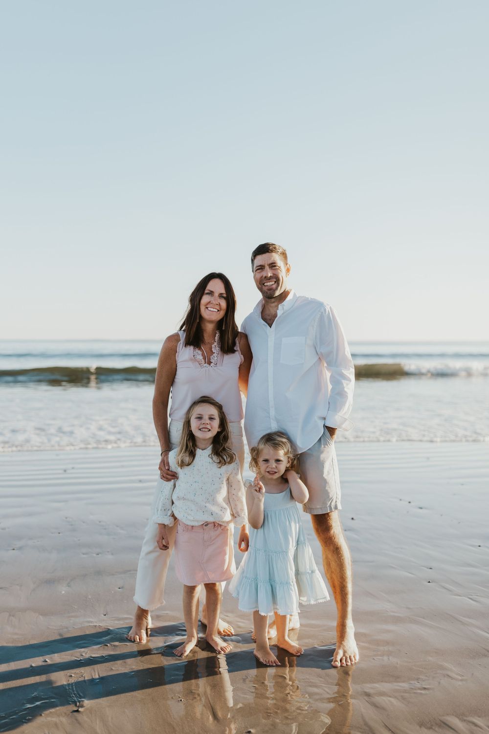 Family of four in white and light colored clothing posing together on a beach at sunset.
