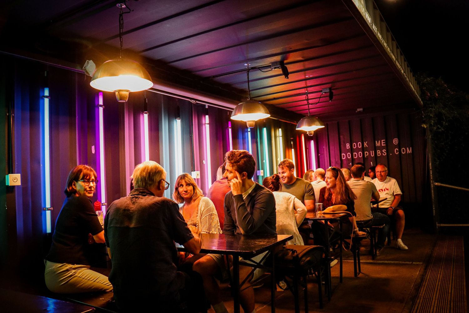 People gather in a dimly lit hallway illuminated by vibrant neon lights and colored tubes creating a moody atmosphere.