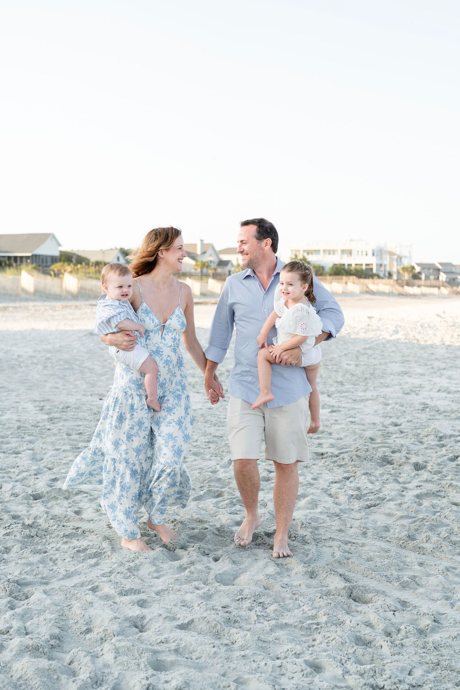 A family walks together on a sandy beach at sunset in coordinated light-colored outfits.