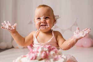 Baby covered in cake frosting smiles and throws hands up during first birthday cake smash photo session.