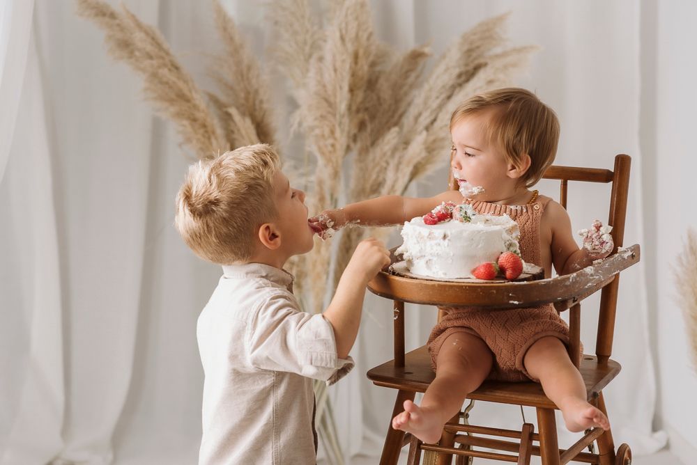 Two small children in white outfits share a moment near high chair with pampas grass decor in background.