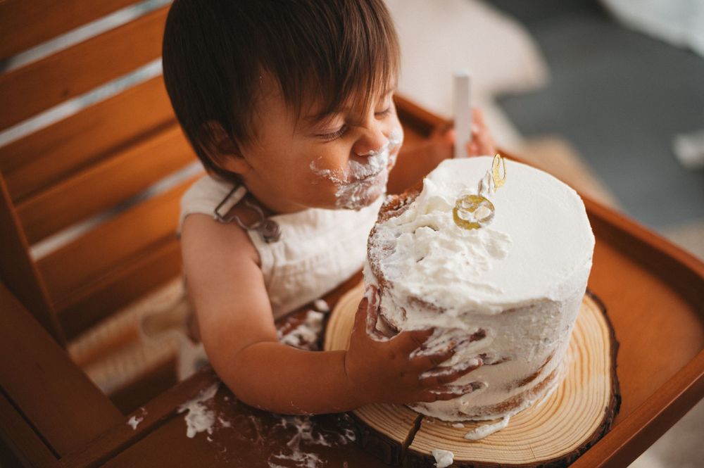 Toddler plays with a white frosted cake while sitting on wooden furniture.