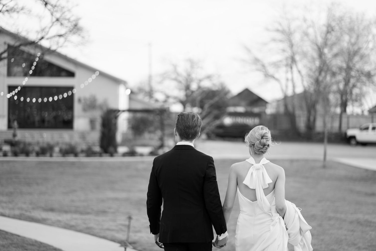 Black and white photo of newlyweds walking away on lawn with string lights visible.