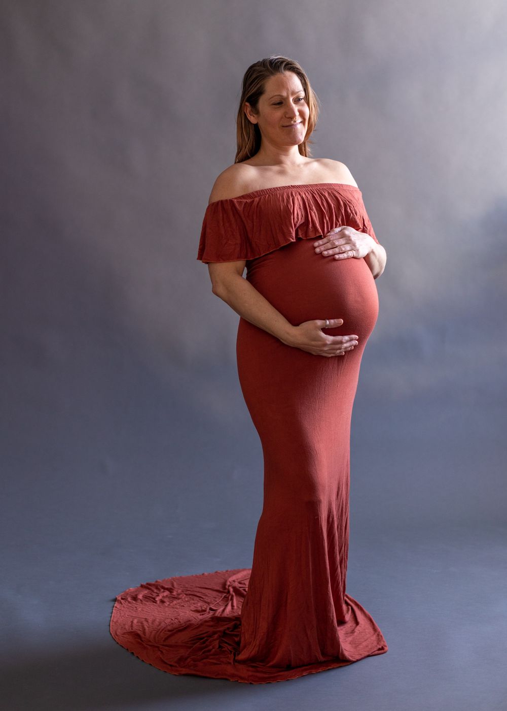 Maternity portrait featuring a rust-colored off-shoulder dress against dramatic cloudy backdrop.