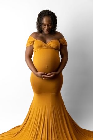 Expectant mother in mustard yellow dress holding her belly against white background.