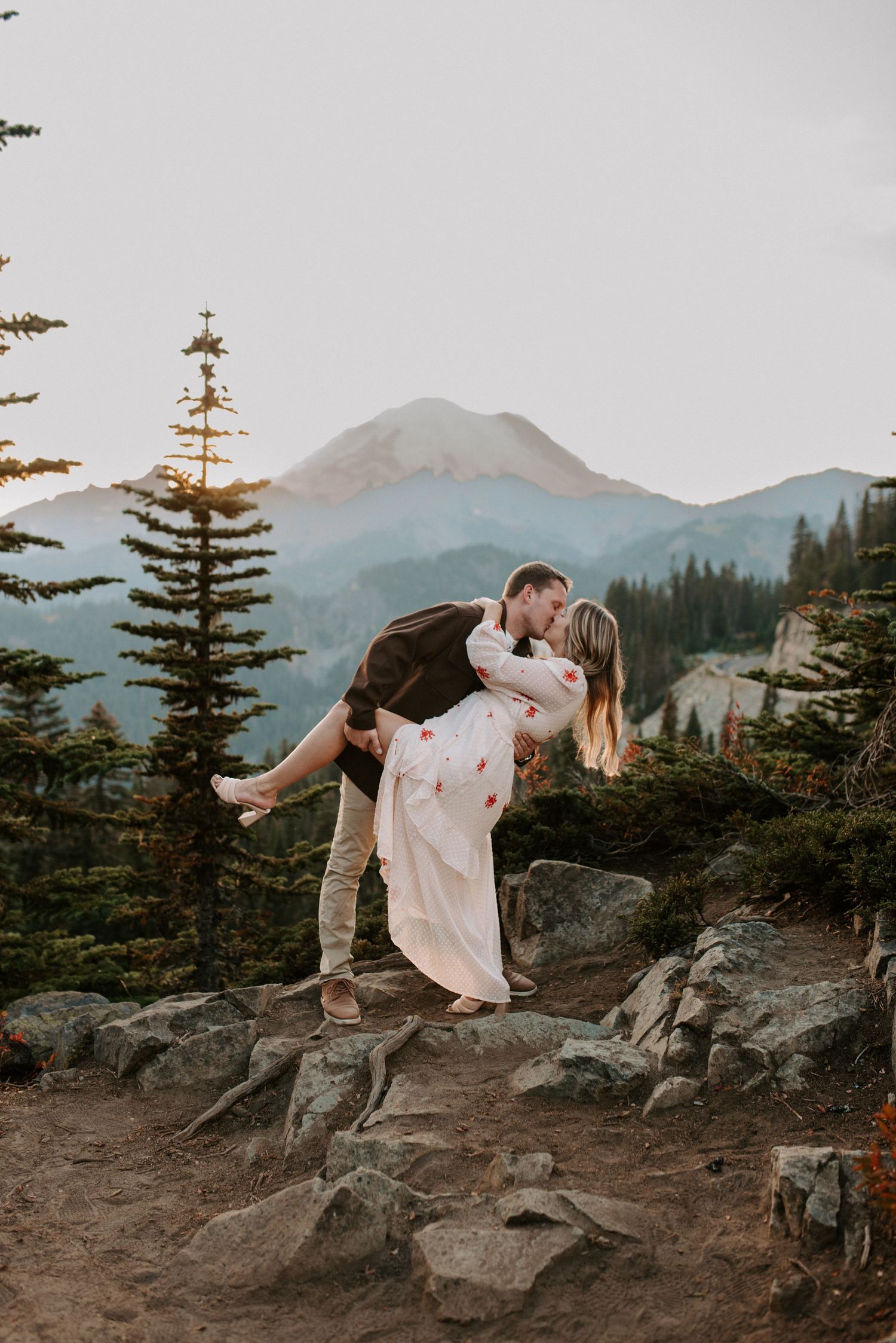 Romantic couple poses on rocky mountain overlook during sunset with evergreen trees.