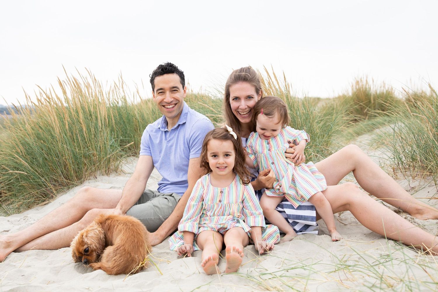 Family relaxing on sandy beach dunes with their dog on a bright day.