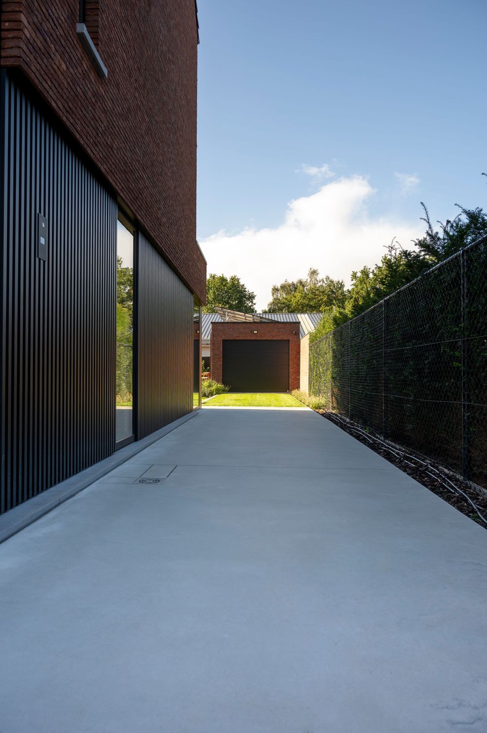 Minimalist concrete pathway bordered by dark vertical panels and hedges against a brick building.