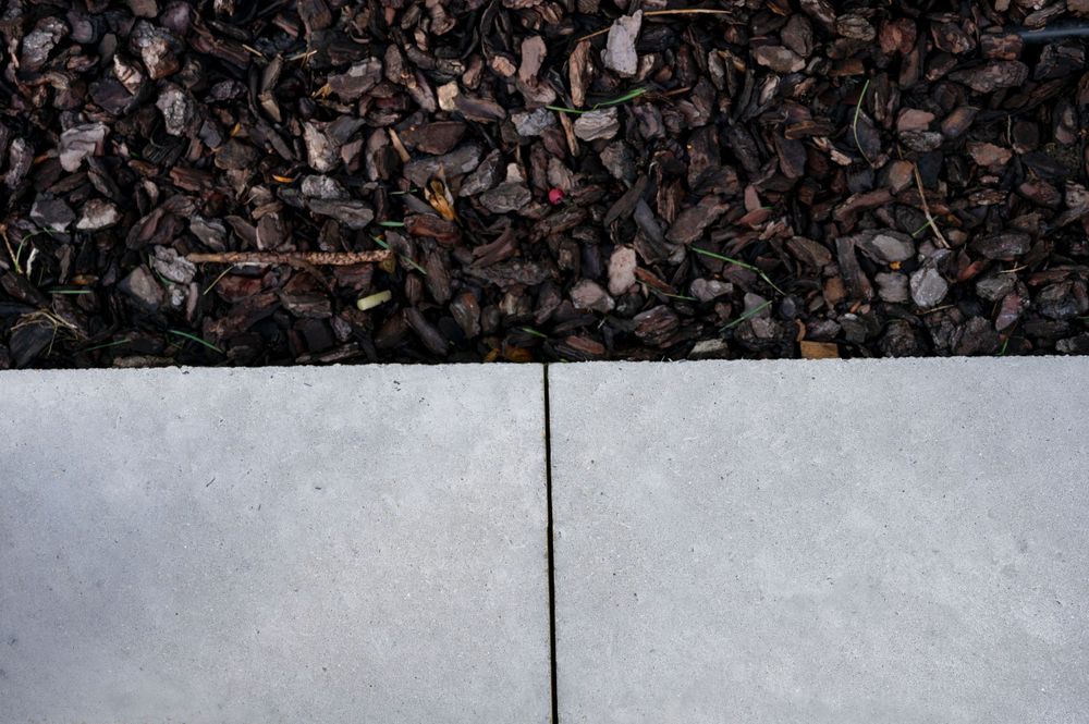 Close-up of concrete pavement joint with dark mulch or stones visible along the edge.