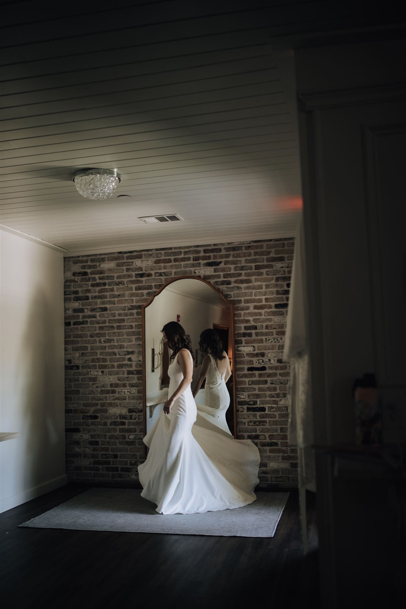 A bride in a white wedding gown admires her reflection in an arched mirror against a brick wall interior.