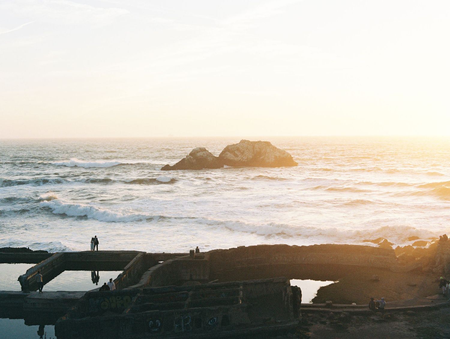 Sunset over rocky coastline with ocean waves crashing against dark silhouetted rocks.