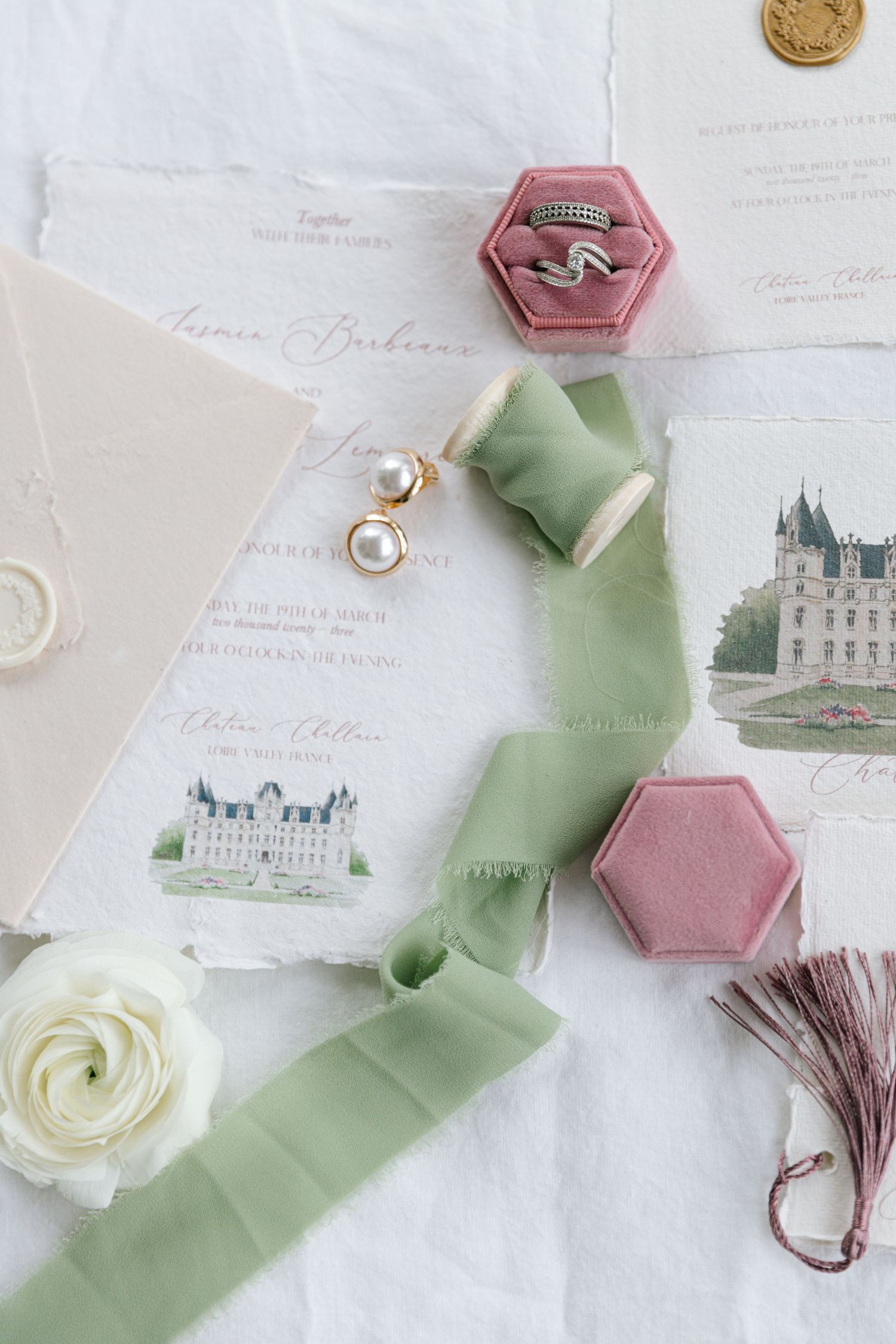 Elegant wedding details including a pink hexagon box, pearl earrings, green silk ribbon, and castle illustrations on paper.