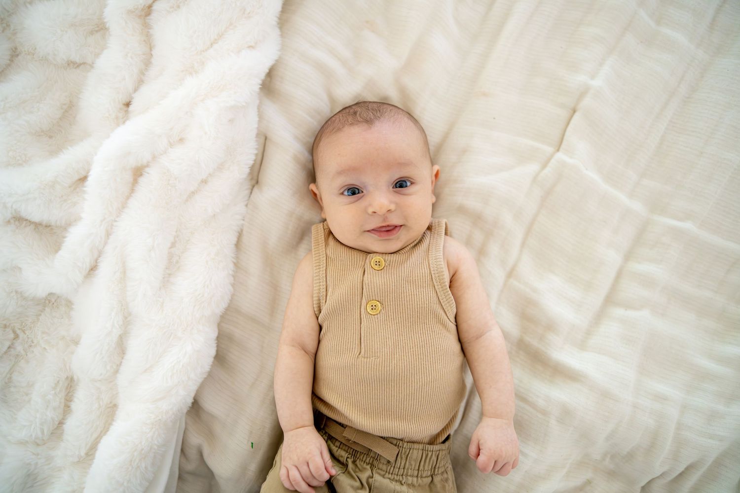 Baby in tan onesie lying peacefully on soft white blanket.