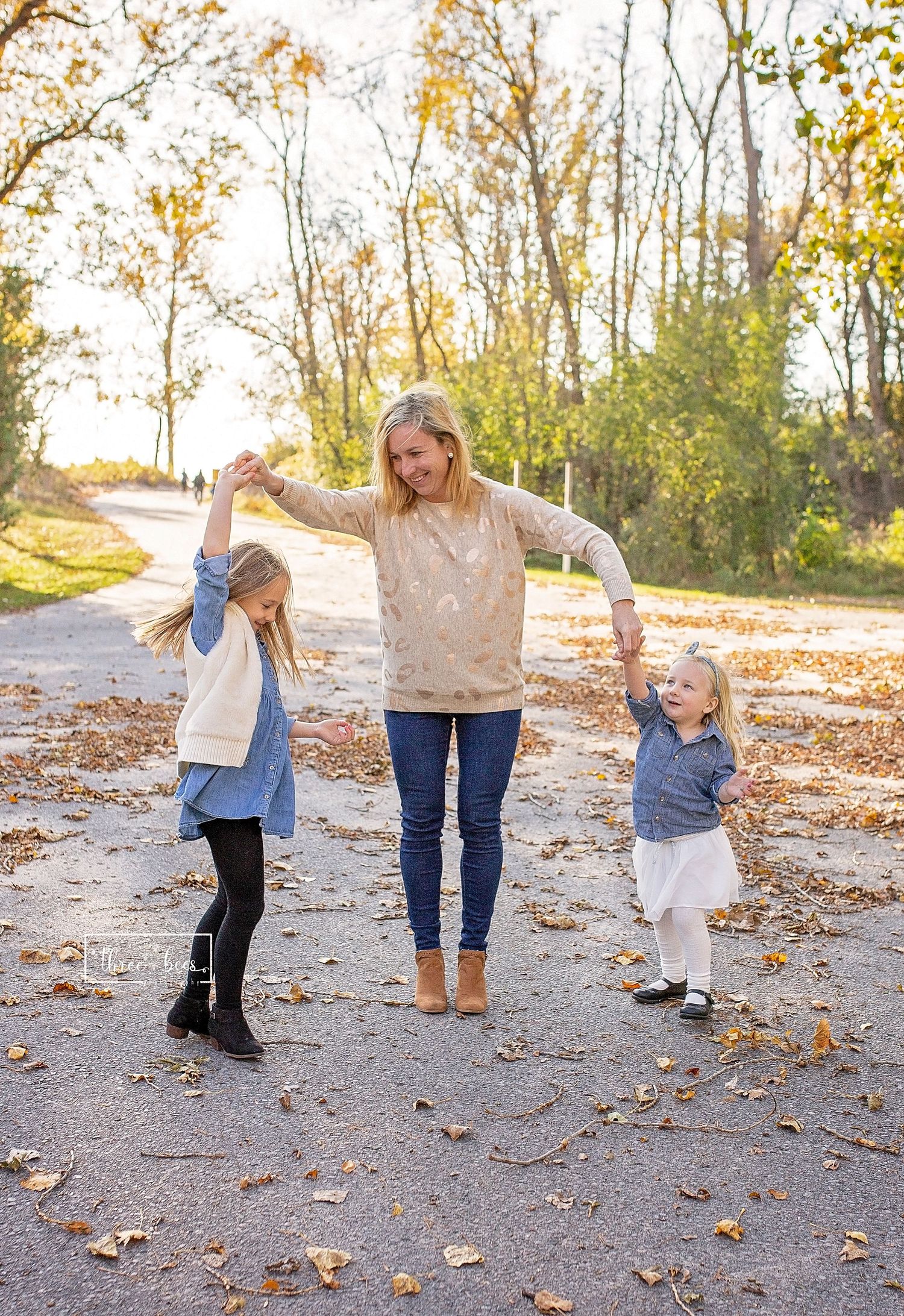 Three people dancing and playing in fallen autumn leaves on a sunny path.