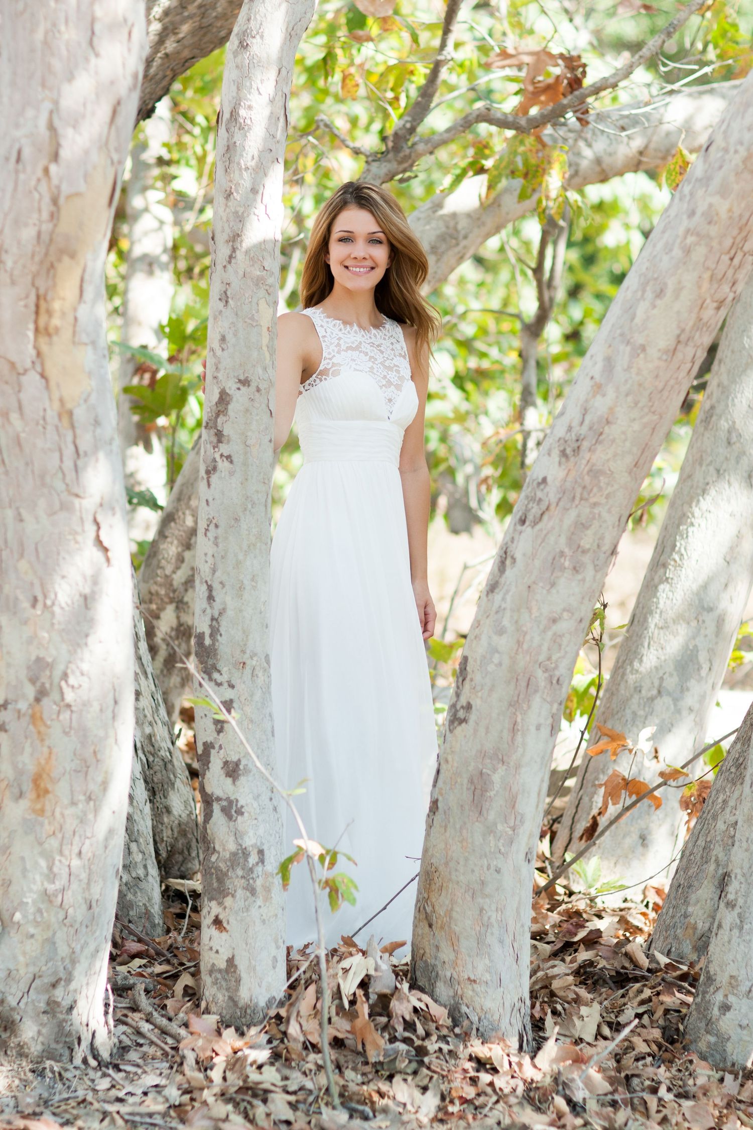 White wedding dress photographed among tree trunks in natural outdoor setting.
