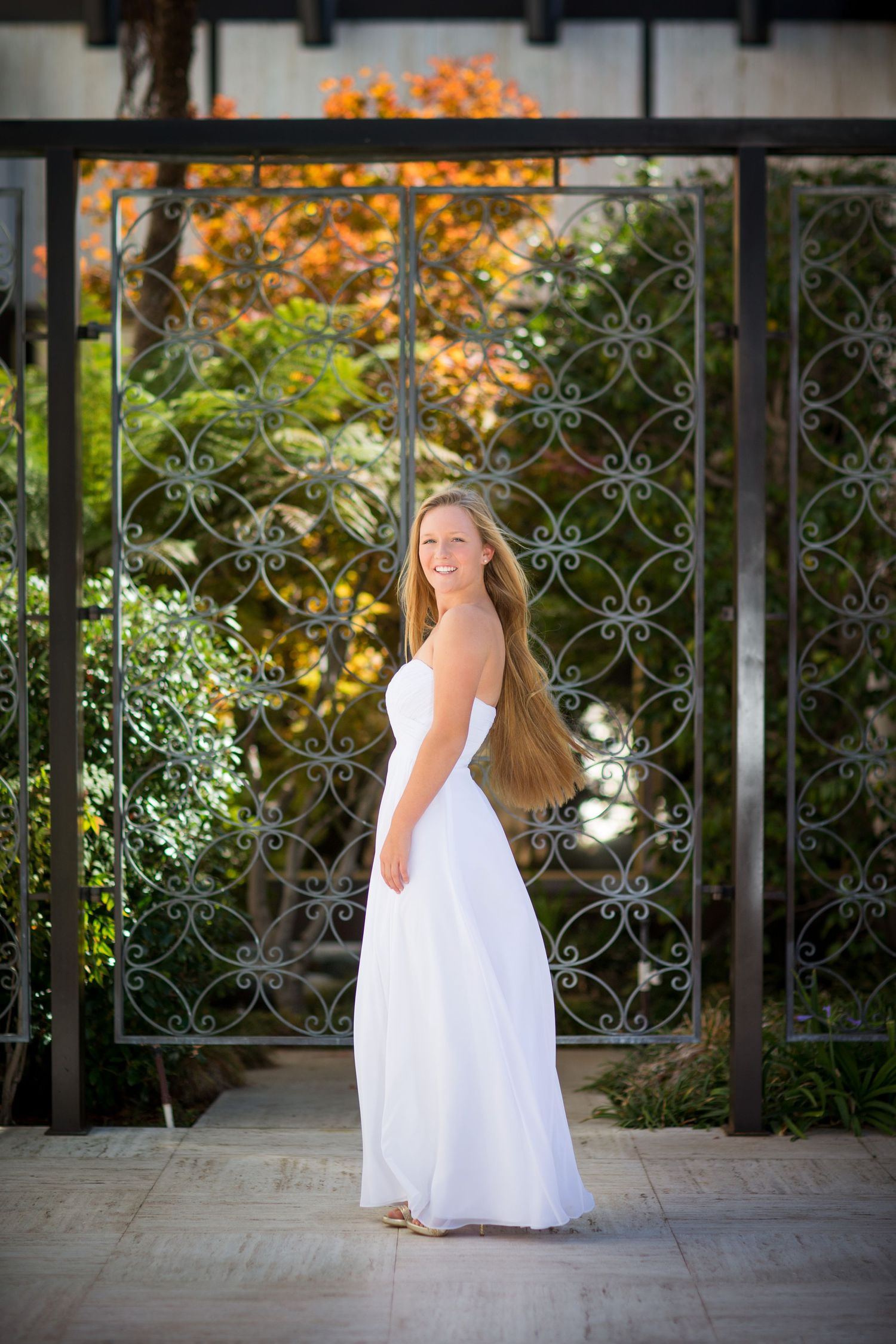 Bride in flowing white gown poses by ornate garden gate with autumn foliage backdrop.