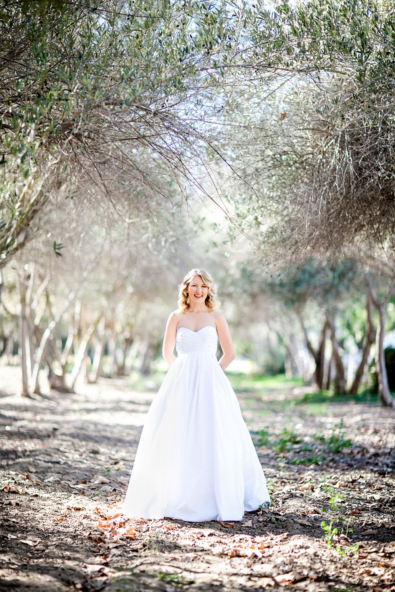 A white wedding gown glows in natural light along a tree-lined path in an outdoor setting.