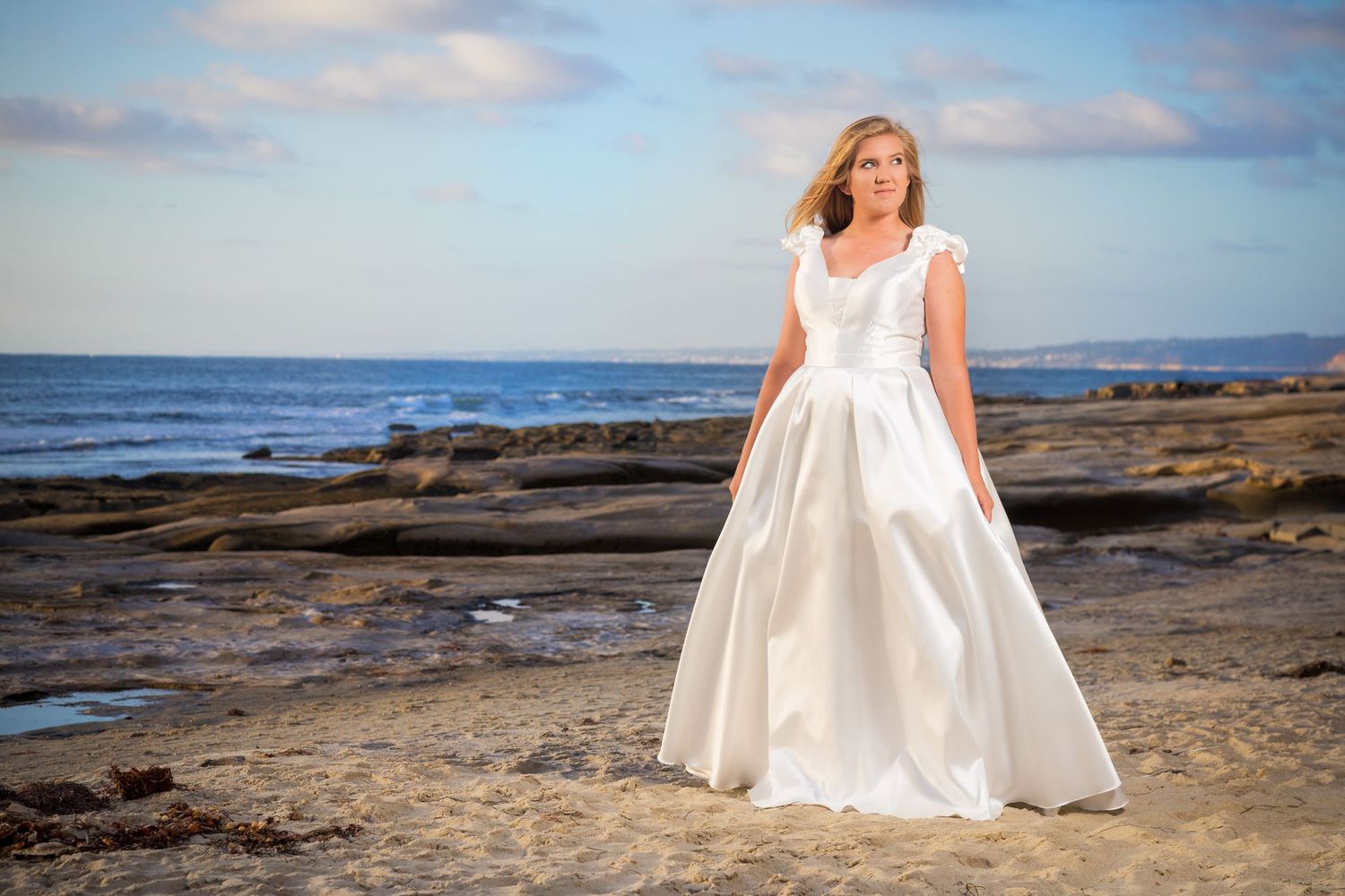 A classic white wedding gown photographed on rocky coastal shores at sunset.