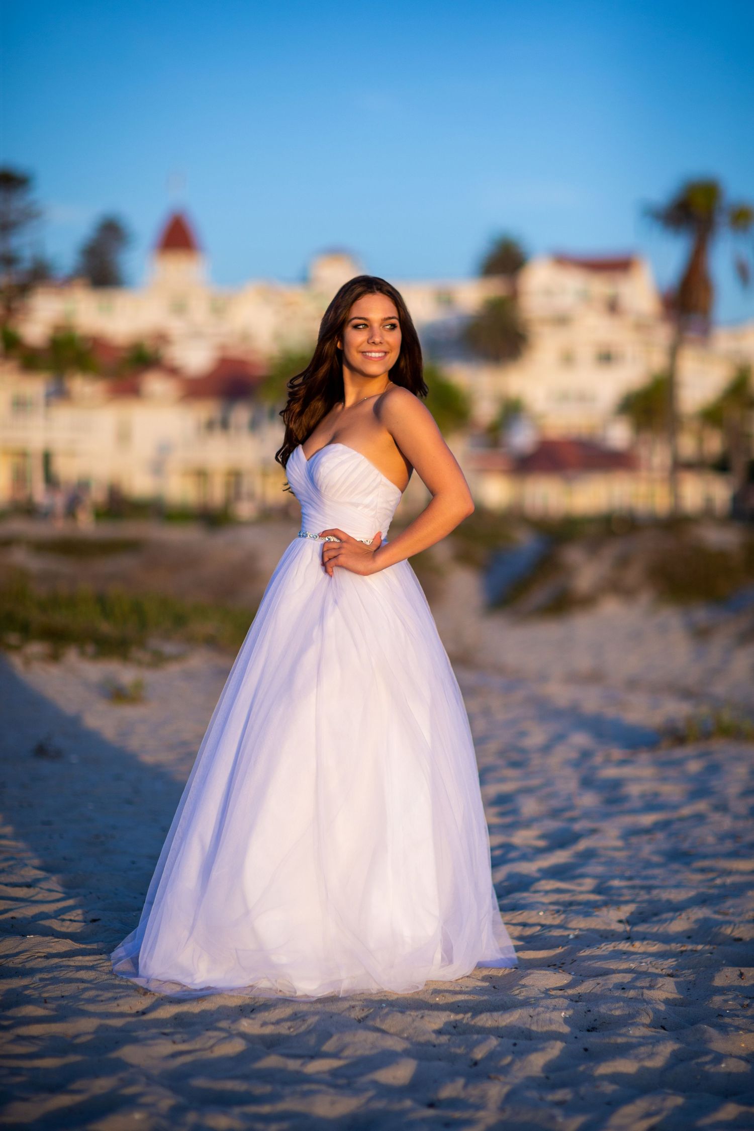 Woman in white ball gown poses on beach with historic buildings in background.
