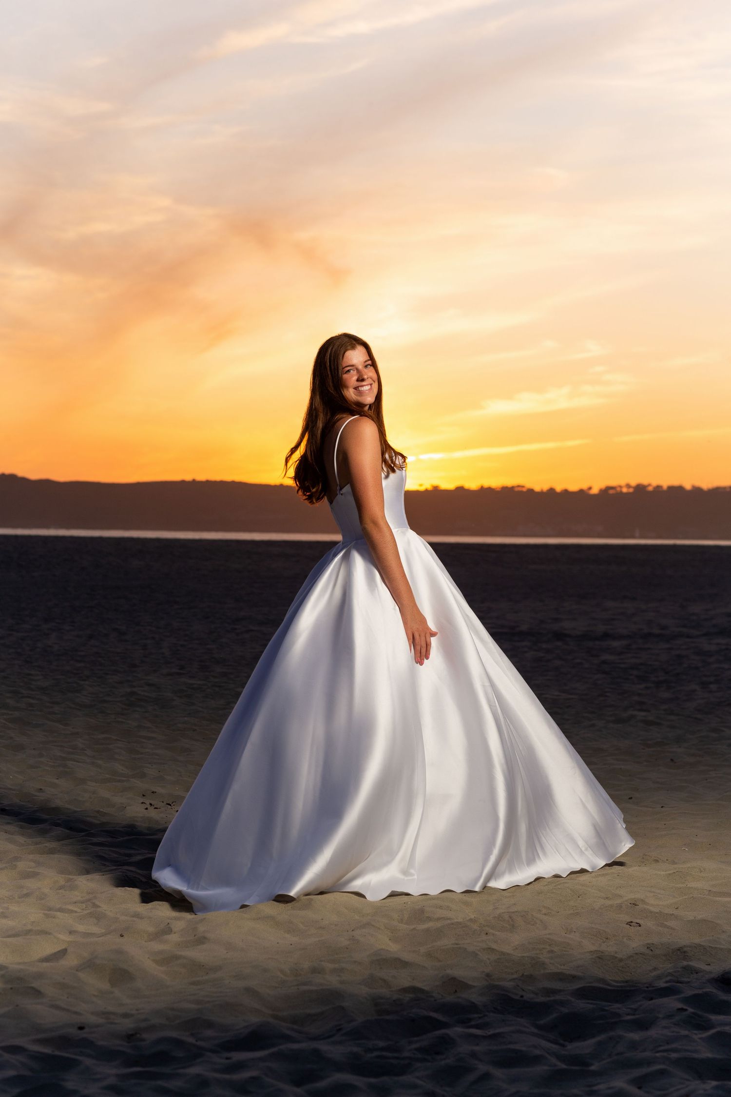A bride in a white ball gown poses against a dramatic sunset on a beach.