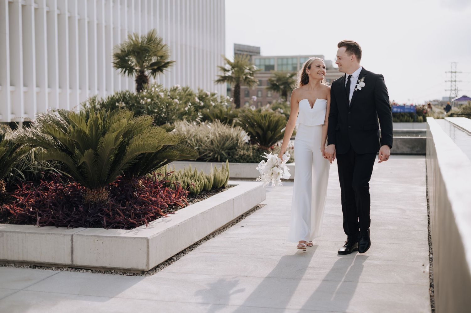 Couple walking together along a landscaped pathway in wedding attire.
