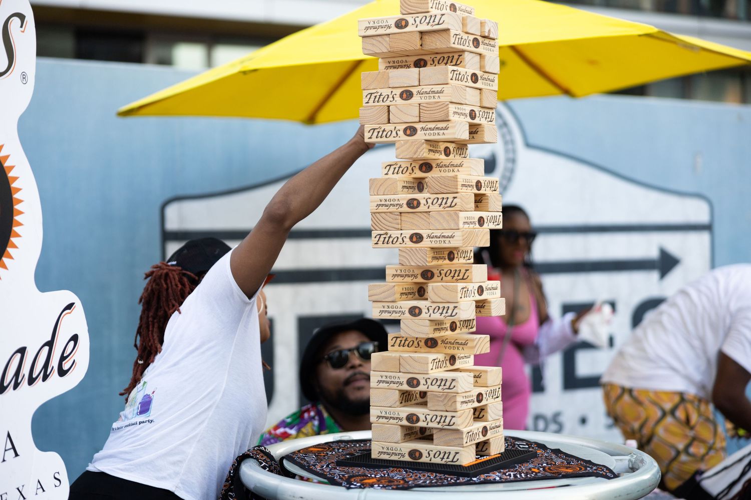People playing a giant Jenga game at an outdoor festival under a yellow umbrella.