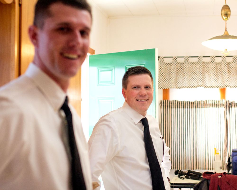 Two men in formal attire smiling in a room with beige curtains and pendant lighting.
