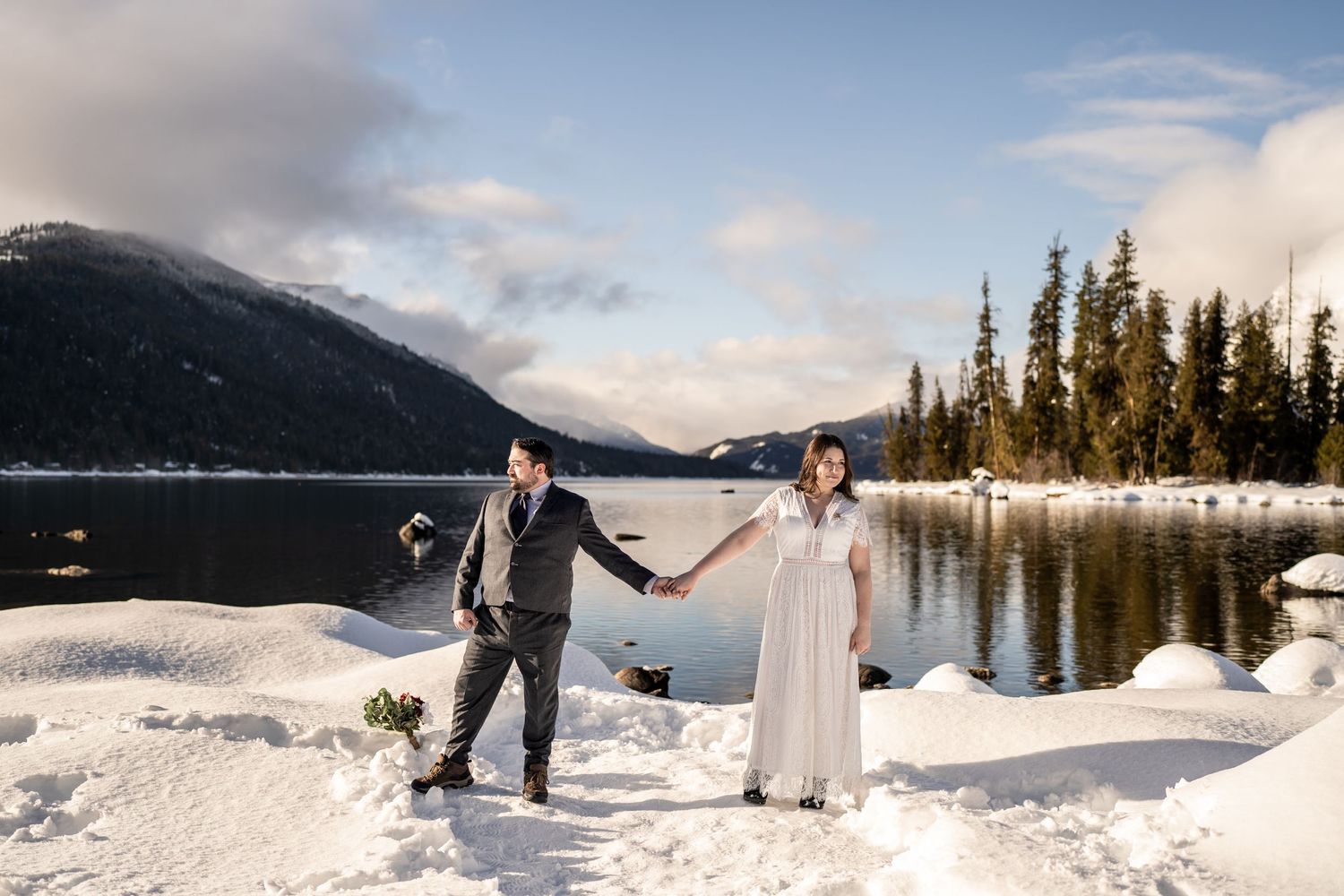 A couple holding hands in winter wedding attire against a snowy mountain lake landscape.