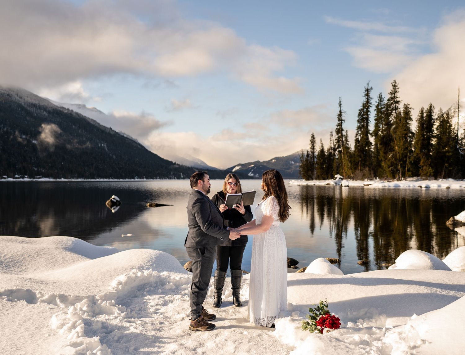 A winter wedding ceremony takes place on a snowy lakeshore with mountain and forest views.