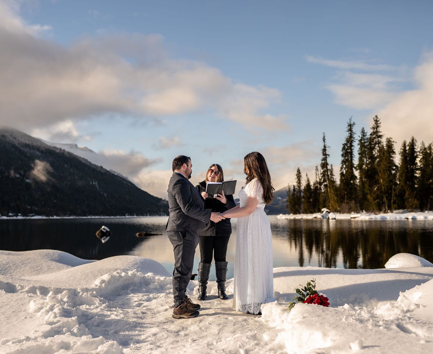 Wedding ceremony unfolding beside a serene mountain lake surrounded by snow and evergreen trees.