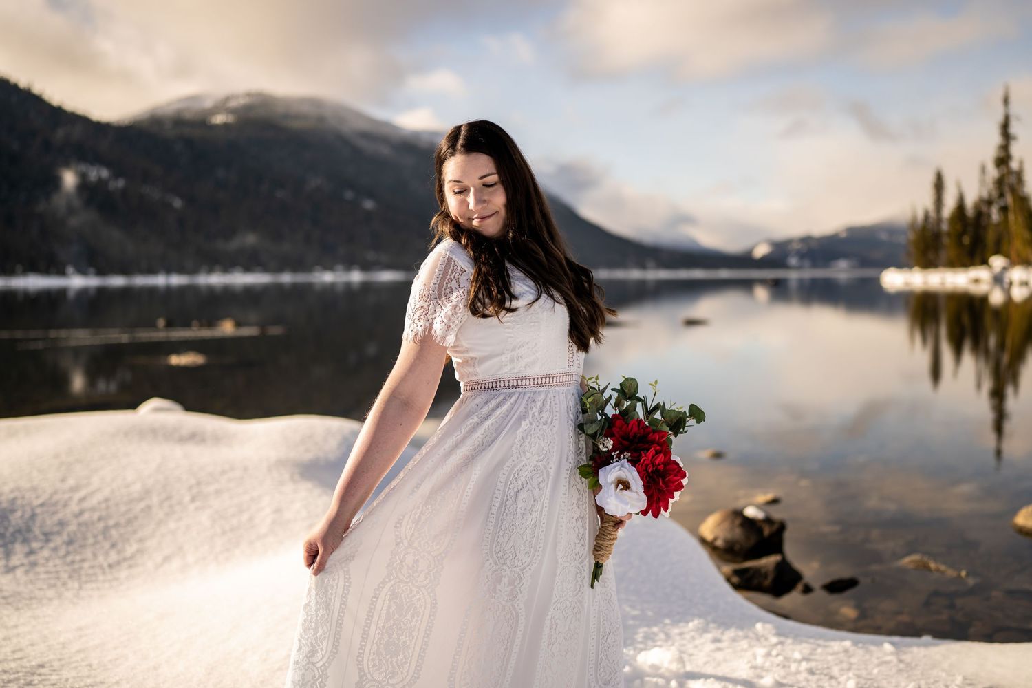 A bride in a flowing white dress holds a red bouquet against a snowy mountain lake backdrop.