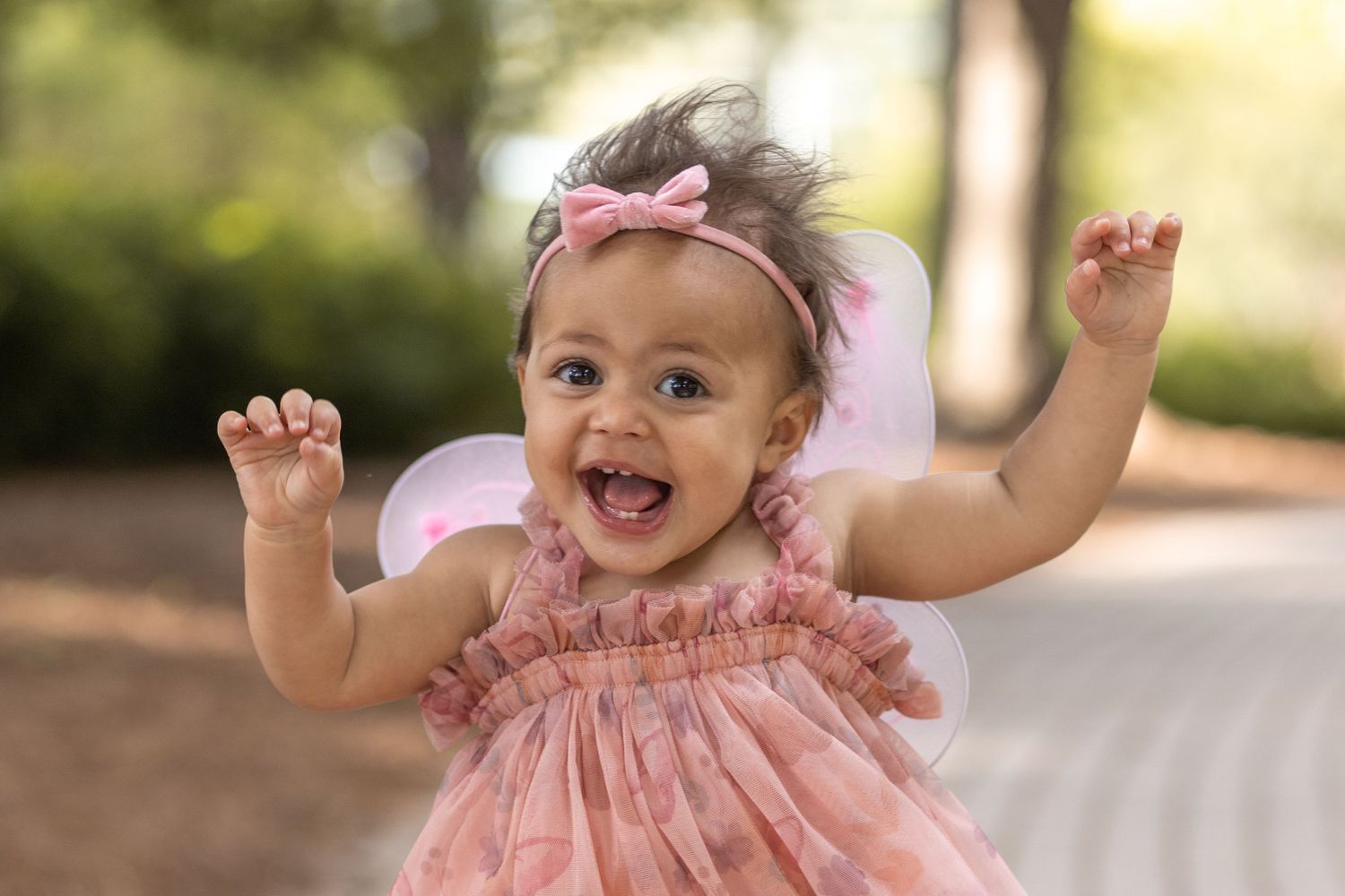 A smiling 1 year old girl in a pink tulle dress and headband runs at the camera with her arms up.